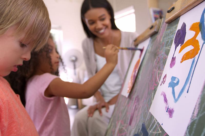 two children painting on an easel as teacher watches