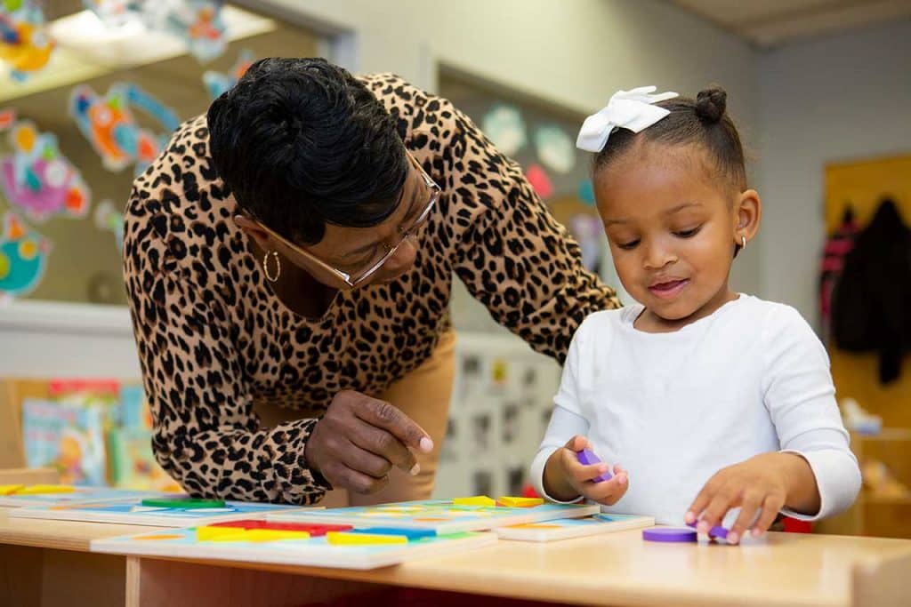 Educator and child in classroom. Child puts shape blocks onto table as educator looks on.