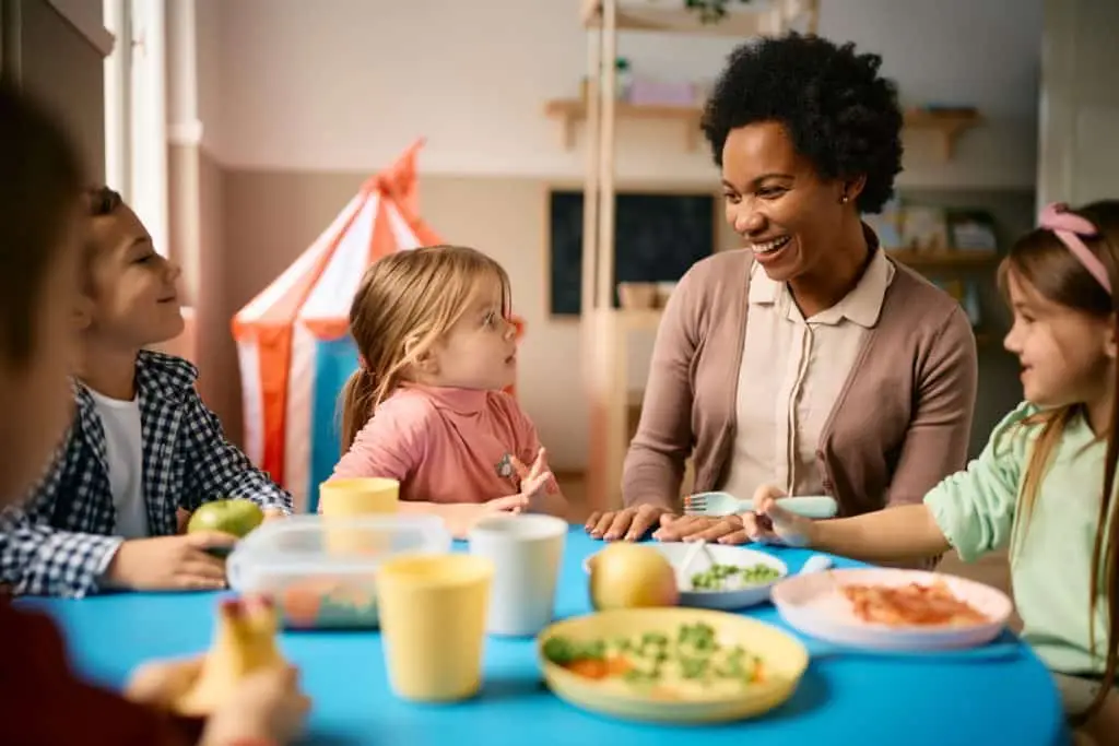 Happy teacher talking to group of kids during lunch time at preschool